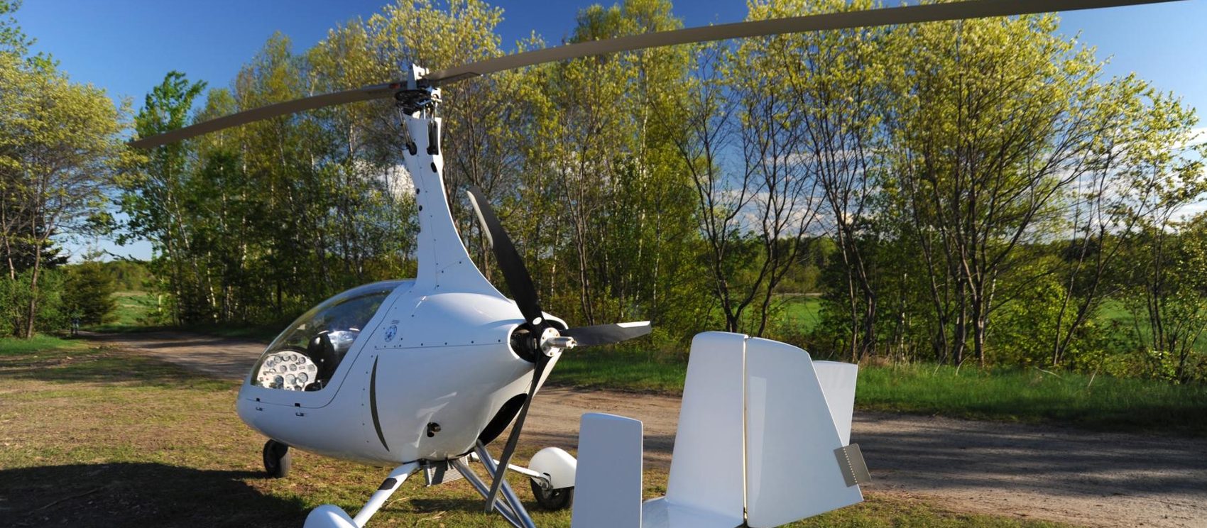White open-frame gyroplane parked on a gravel track in woodland
