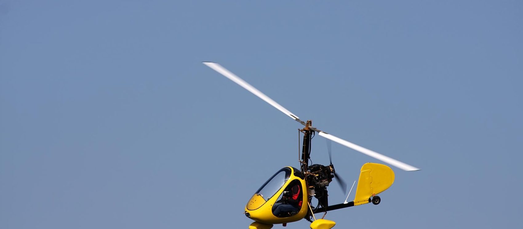 Yellow enclosed-cabin gyroplane in flight against a clear blue sky